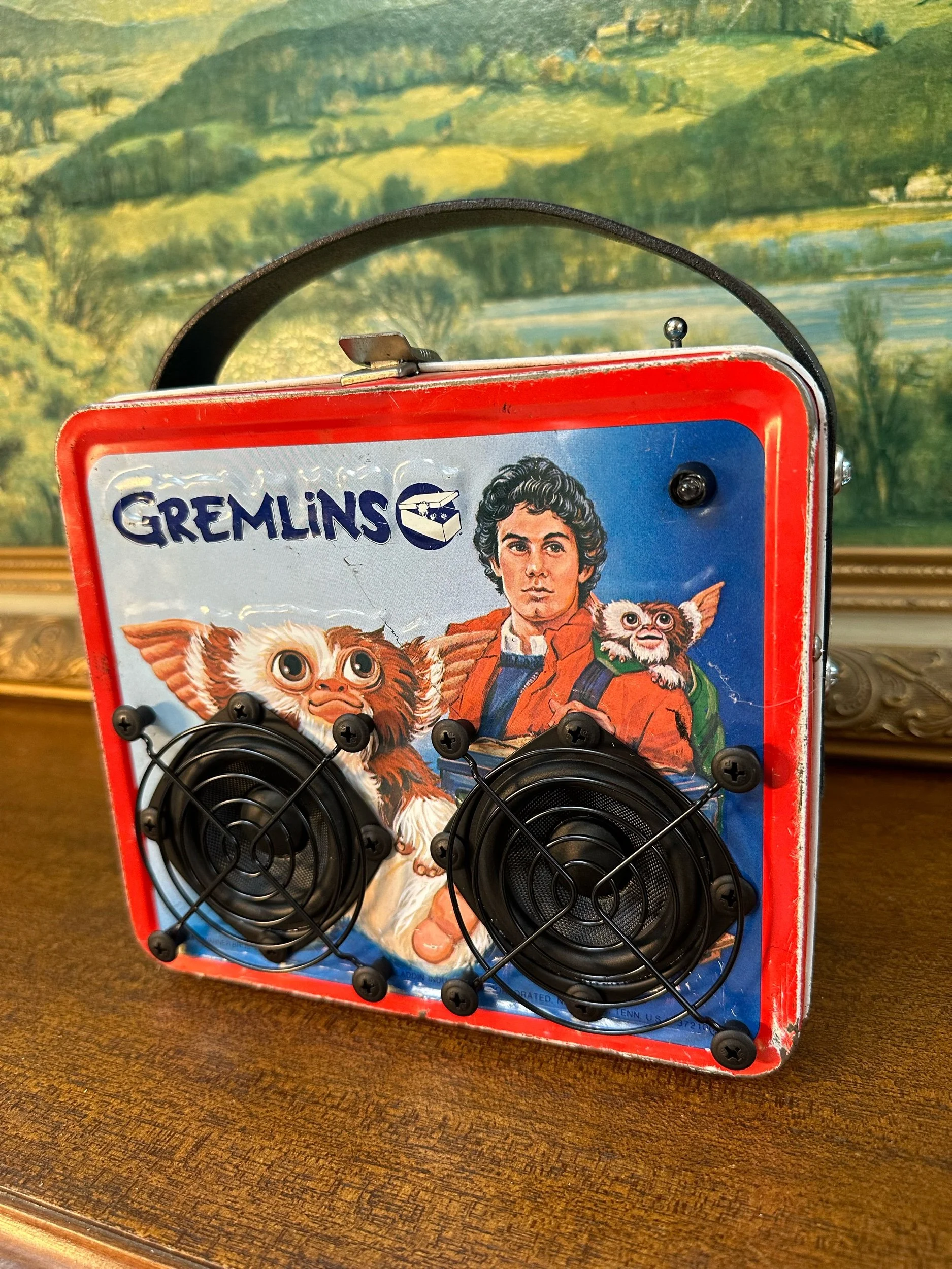 Vintage Gremlins lunchbox converted into a speaker with an illustration of movie characters, featuring two speaker grills and a leather handle, set against a scenic painting background.