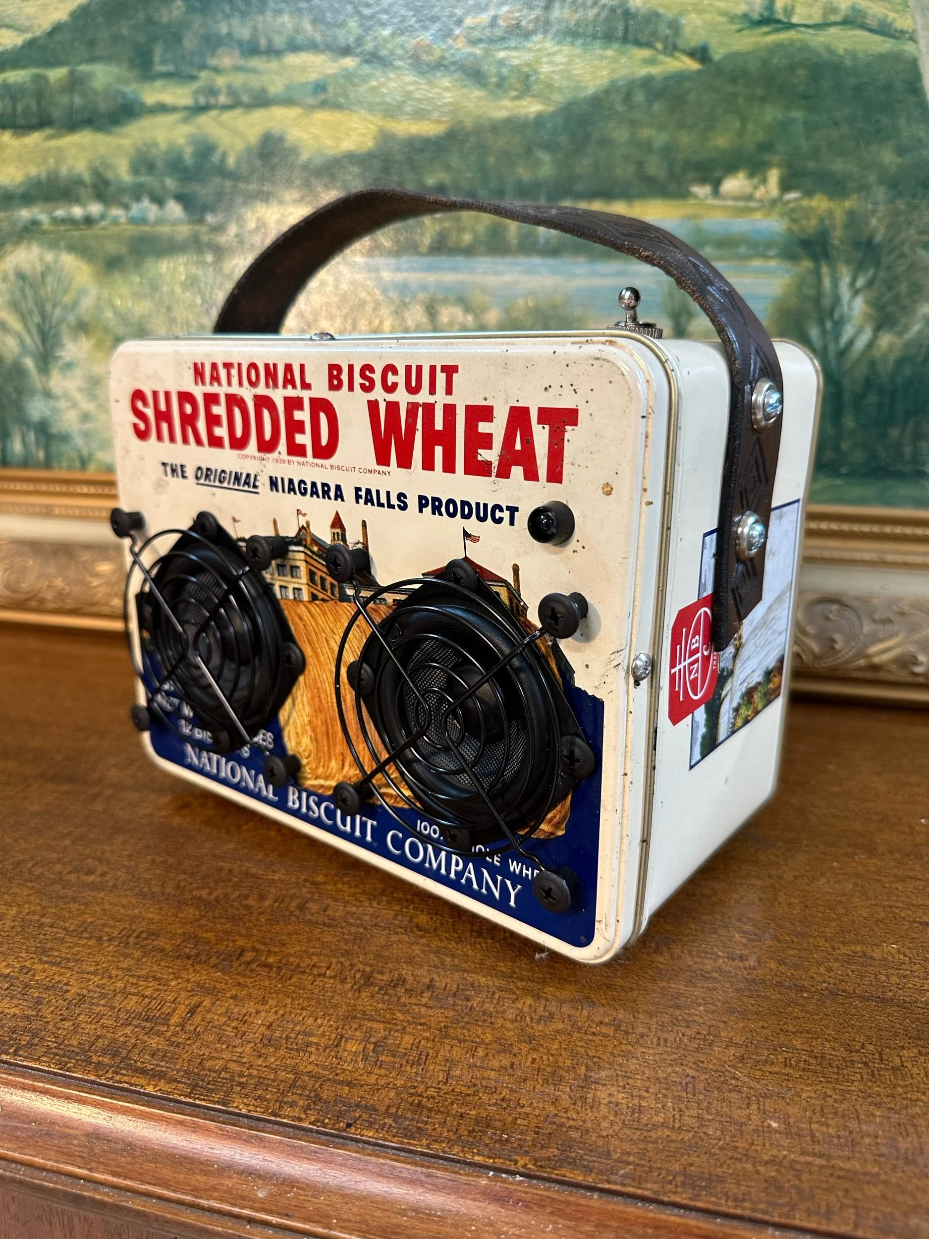 Vintage National Biscuit Company tin with a retro Shredded Wheat advertisement and two industrial fans attached, displayed on a wooden surface with a scenic backdrop.