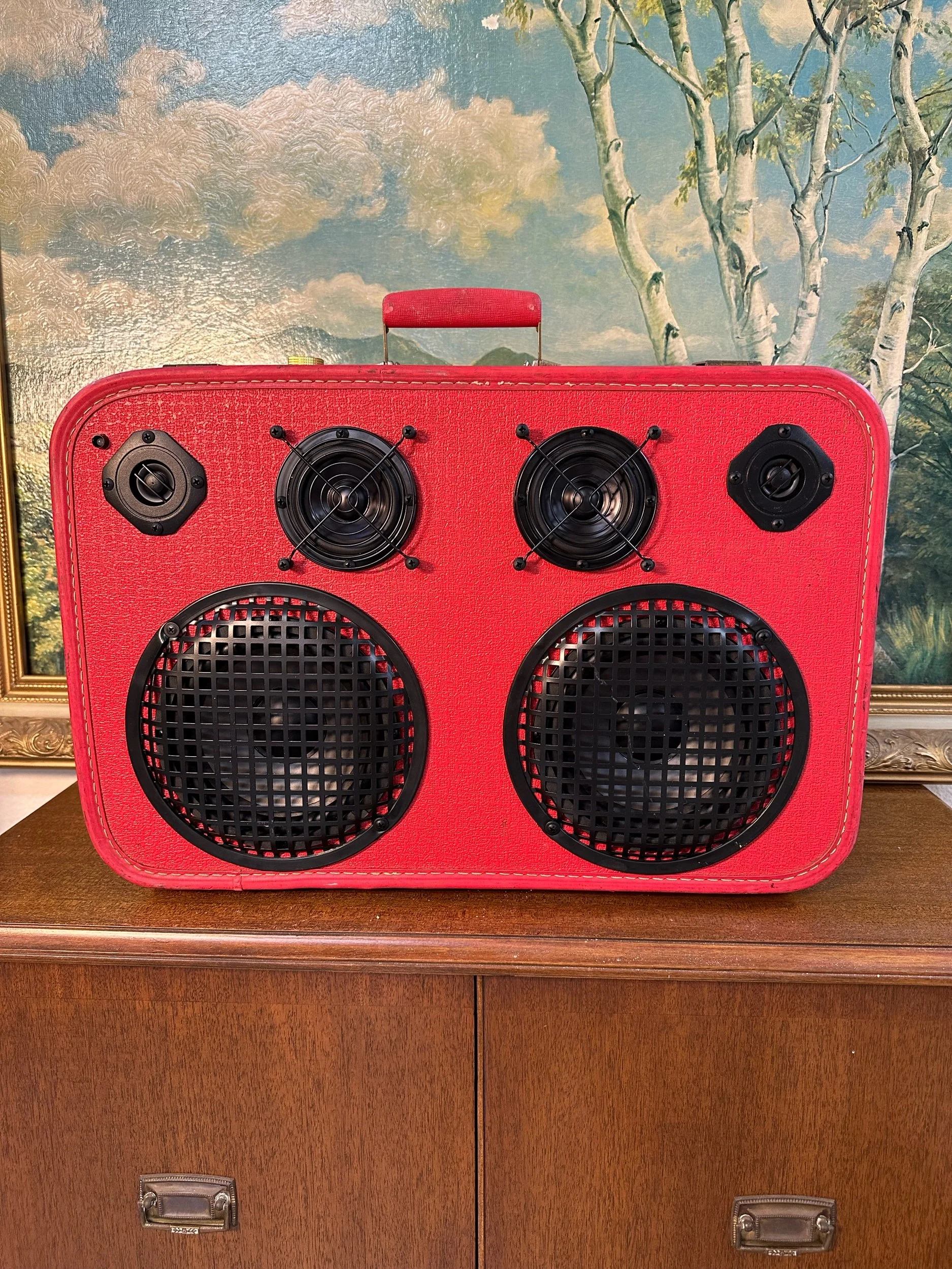 Red vintage-style suitcase with built-in speakers on a wooden cabinet, decorative painting in the background.