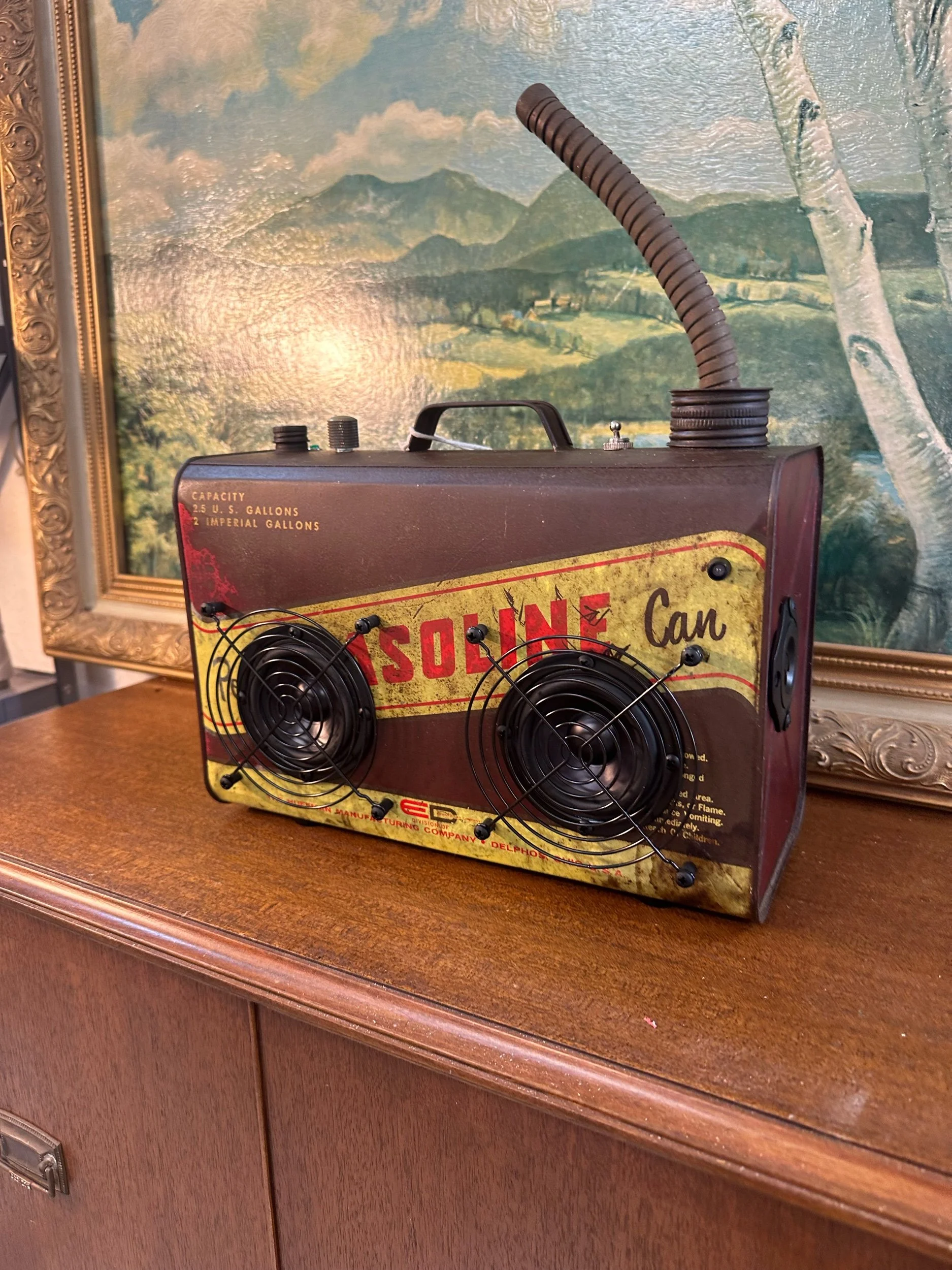 Retro-styled speaker made from repurposed gasoline can with vintage design details, placed on wooden furniture, landscape painting in background.