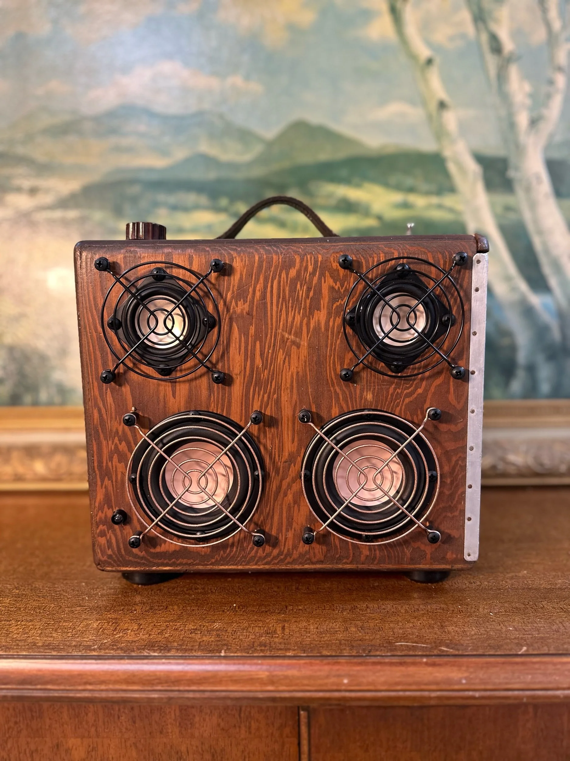 Vintage wooden amplifier with four circular speakers and metal grilles on a wooden surface, in front of a landscape painting.