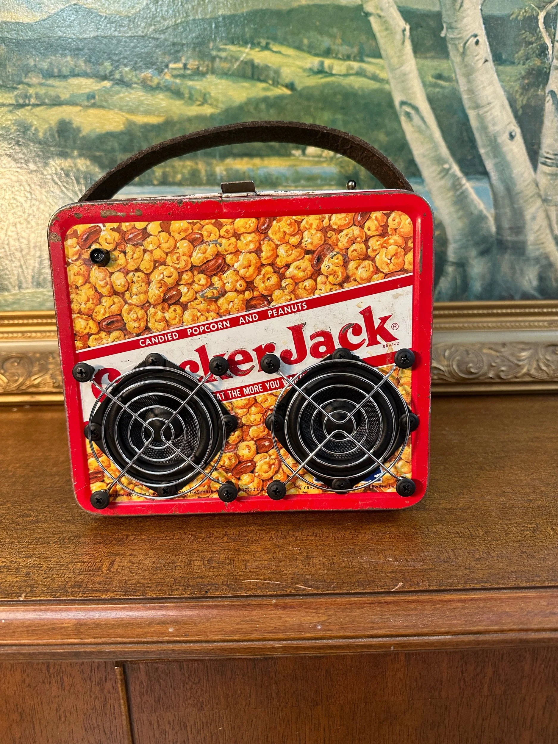 Vintage Cracker Jack tin lunchbox with two speakers attached, displayed on a wooden surface with a landscape painting in the background.