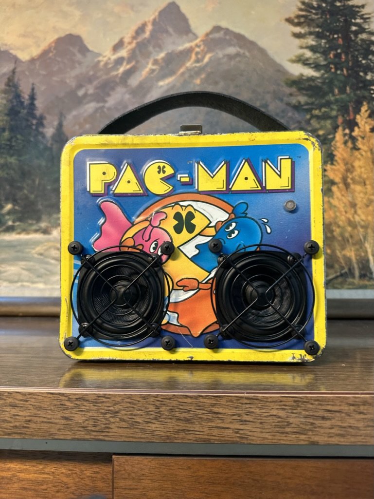 Retro Pac-Man lunchbox repurposed into a speaker with colorful characters on the front, placed on a wooden surface.