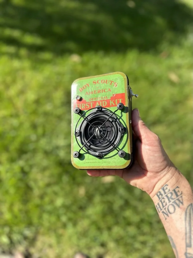 A hand holding a green and red Boy Scouts of America official first aid kit with a circular black wire grid on the front, outdoors with green grass in the background. The person has a tattoo on their forearm.