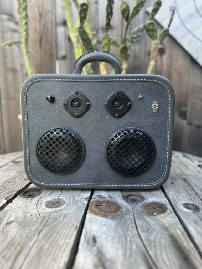 Vintage-style suitcase boombox with speakers and handle sitting on a wooden table, background with cactus and wooden fence.