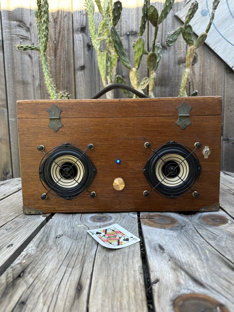 Retro-style speaker box with wooden casing and metal accents, featuring two circular speakers, a central control knob, and a small blue light indicator. A queen of hearts playing card is placed on a wooden surface in front, with cactus plants and a w