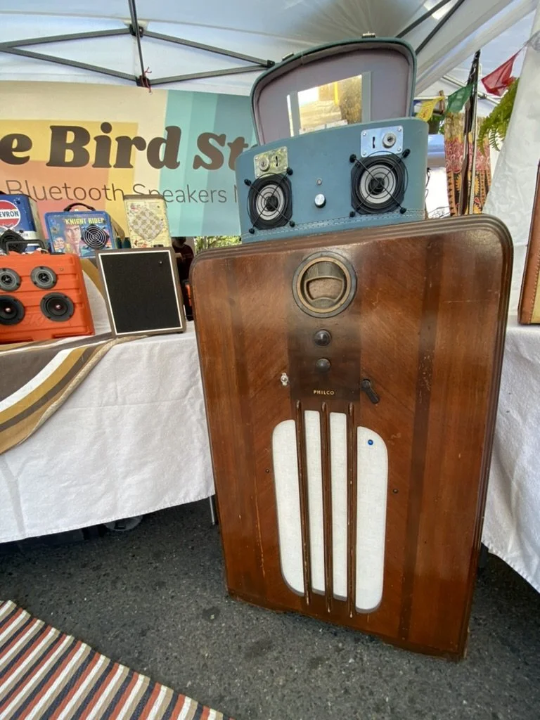 Vintage Philco radio repurposed as a Bluetooth speaker at a market stall, with a sign in the background and various audio equipment displayed nearby.