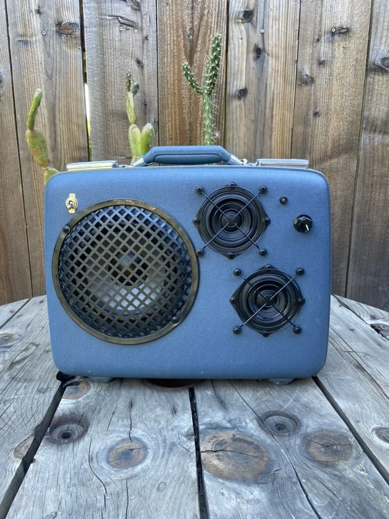 A vintage-style portable speaker housed in a blue suitcase with metal grilles, placed on a wooden surface with a wooden fence and cactus in the background.