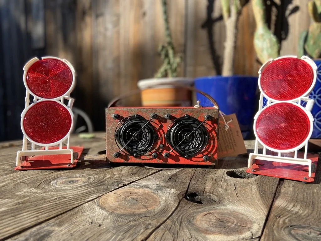 A wooden table with a red-metallic device featuring two fans in the center, flanked by two red reflector warning lights. Background includes potted plants and a wooden fence.