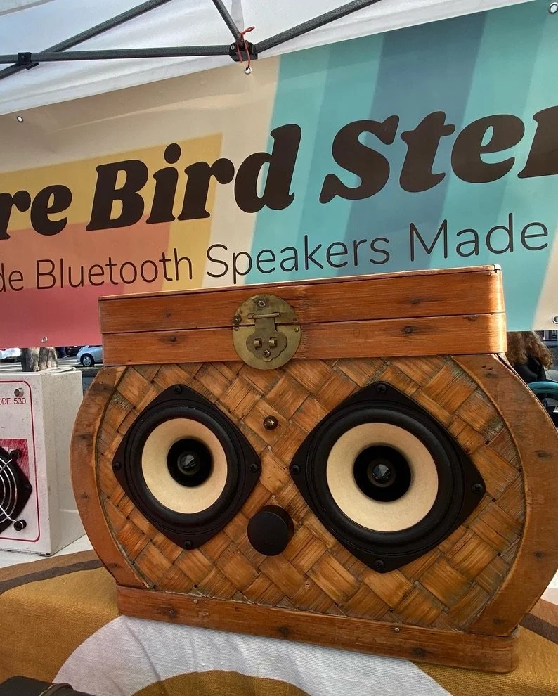 Wooden handcrafted Bluetooth speaker displayed on a table, with a woven design resembling a chest, part of an outdoor market stall with a colorful banner in the background.