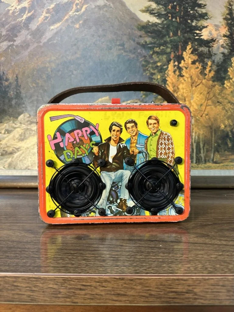 A vintage tin lunchbox featuring a "Happy Days" design with three male characters on the front, retrofitted with speakers, placed on a wooden surface with a scenic mountain painting in the background.