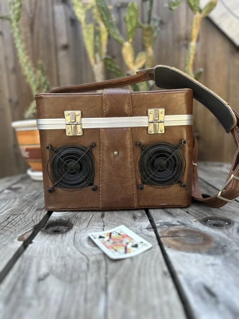 Leather bag with built-in speakers on a wooden table, playing card in foreground, cactus in background.