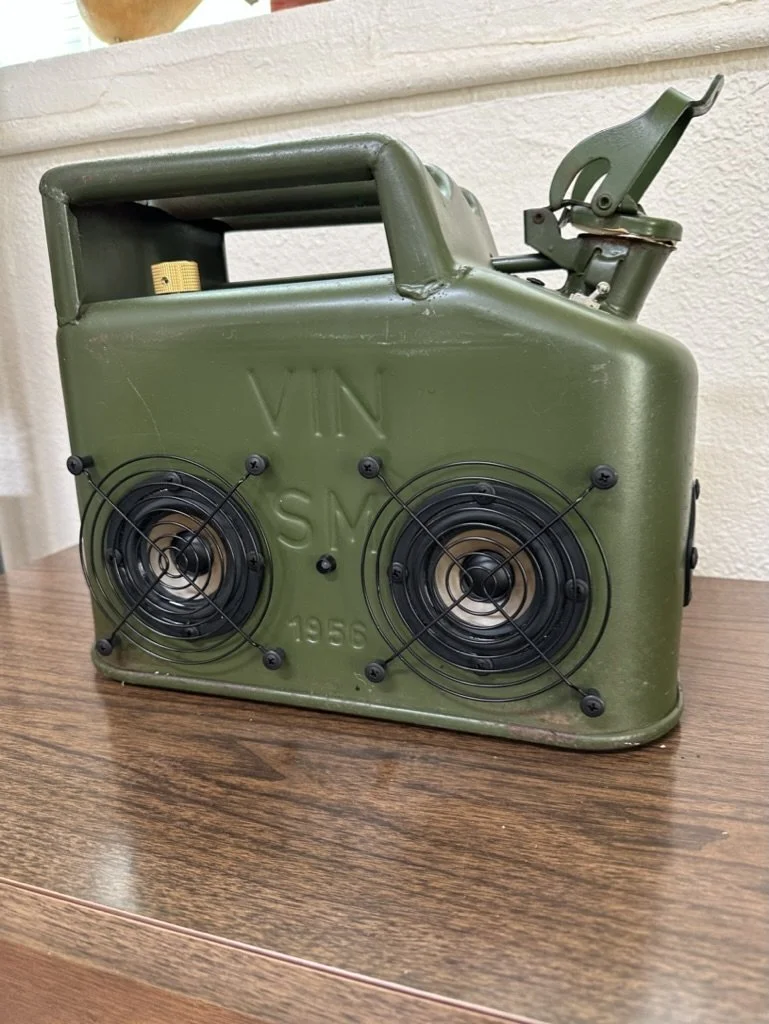 Vintage jerry can repurposed into a portable speaker with two circular speakers mounted on one side, placed on a wooden surface.