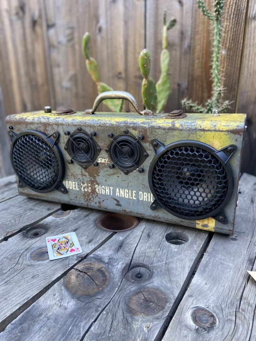 Vintage-style boombox speaker made from an old Model 208 right angle drill case, placed on a rustic wooden table with a Queen of Hearts playing card nearby. Background features a wooden fence and cactus plants.