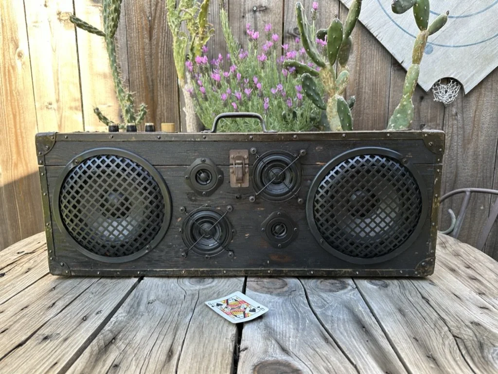 Vintage wooden boombox with large speakers on a rustic table, surrounded by cactus and lavender plants, with a playing card in front.