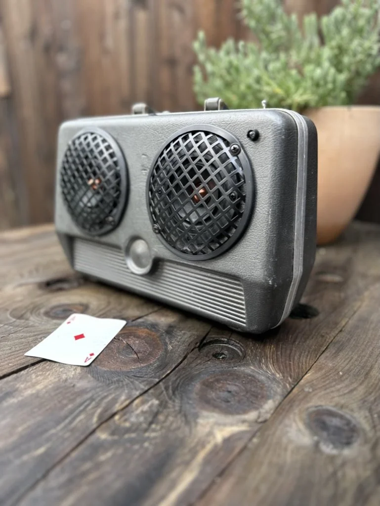 Retro-style radio with two speakers on a wooden surface, potted plant in background, playing card on table.