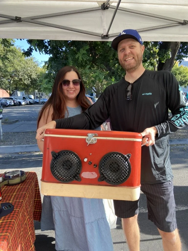Two people smiling, holding a red cooler converted into a stereo speaker, under a canopy at an outdoor market.