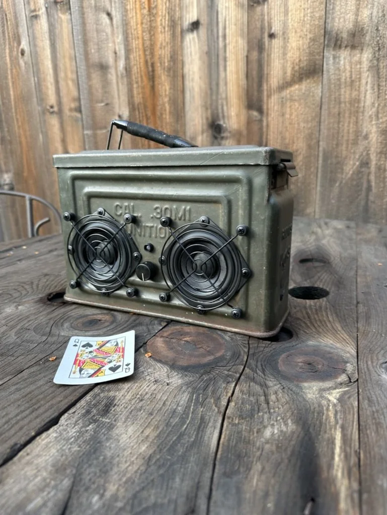 A repurposed ammunition box with two speaker grills, placed on a wooden table next to a playing card.