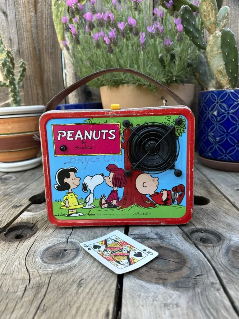 Vintage Peanuts-themed lunchbox with character illustrations, featuring a built-in radio or speaker grill, on a wooden surface with a deck of playing cards. Potted plants with flowers and cacti are in the background.