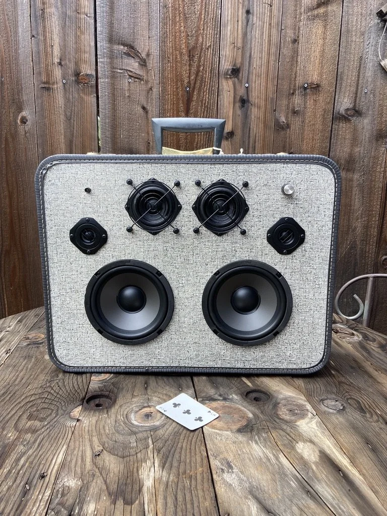 Vintage suitcase converted into a speaker with multiple audio components and a playing card on a wooden surface.