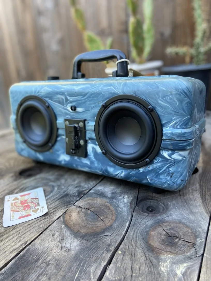 Portable Bluetooth speaker with two large circular speakers and a marble-patterned blue casing, placed on a wooden surface. A playing card is lying on the table next to the speaker.