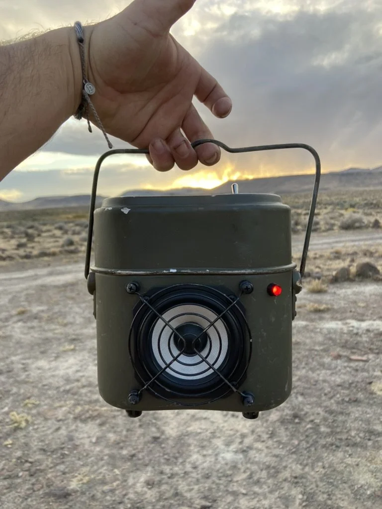 Person holding a portable green speaker outdoors with a sunset in the background.