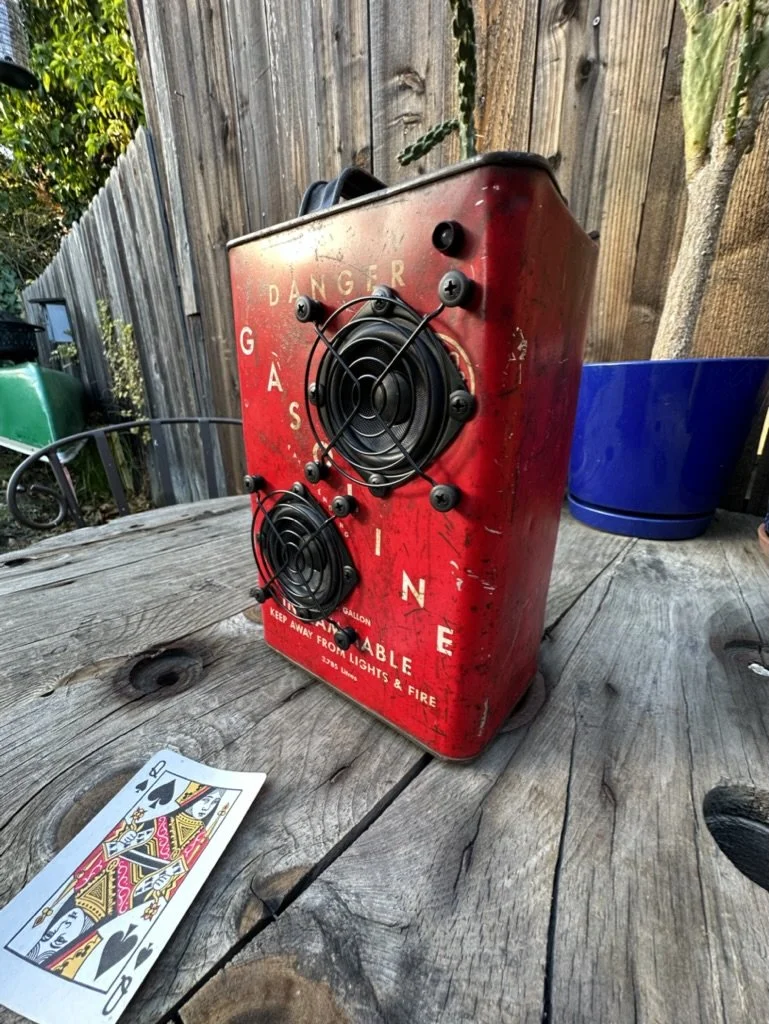 Red vintage gasoline can repurposed with speakers on a wooden outdoor table, next to a playing card, with plants and a wooden fence in the background.
