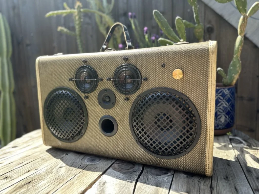 Vintage-style speaker with multiple drivers, placed outdoor on a wooden surface, surrounded by plants.