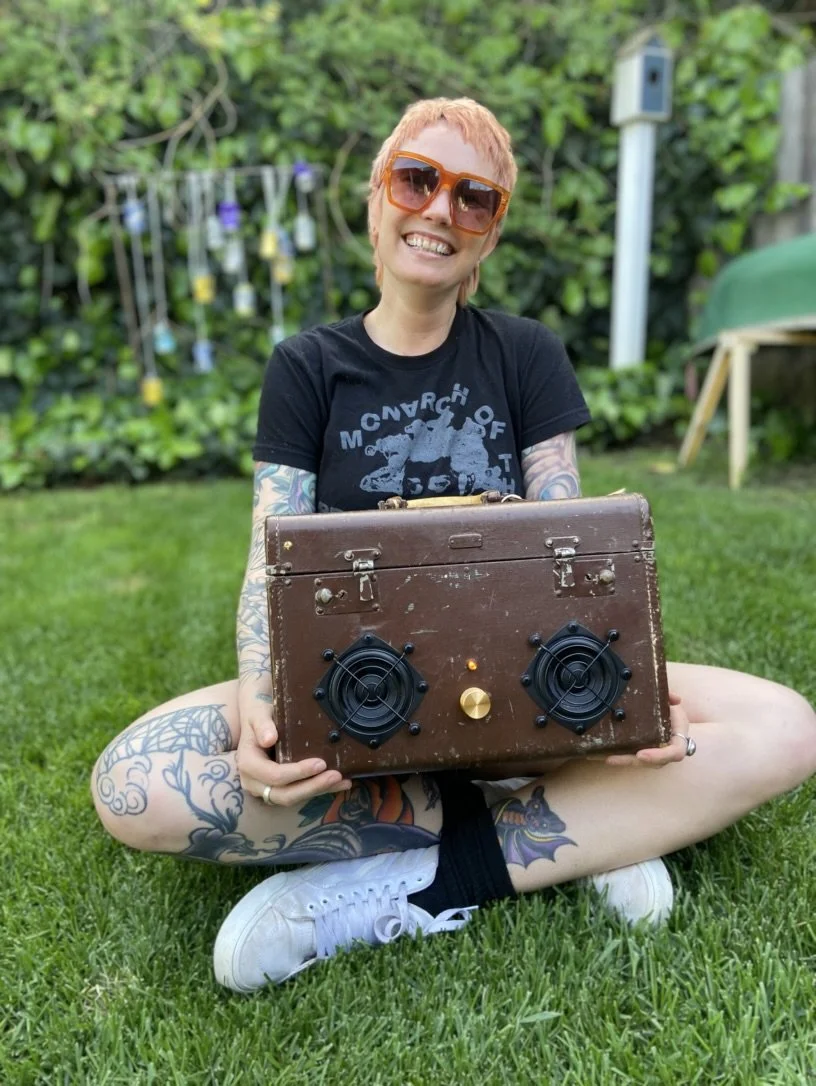 Smiling person with short hair and large sunglasses holding a vintage-looking speaker outdoors on grass, surrounded by greenery.