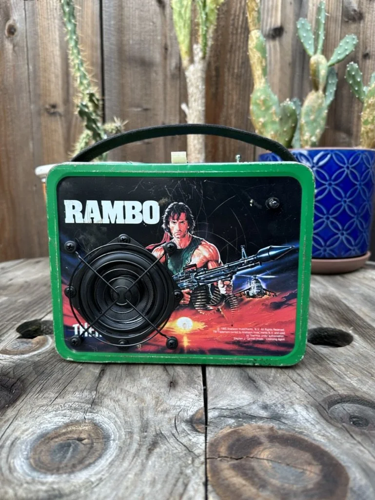 Vintage Rambo lunchbox with speaker featuring movie artwork, on a wood surface with a cactus plant in the background.