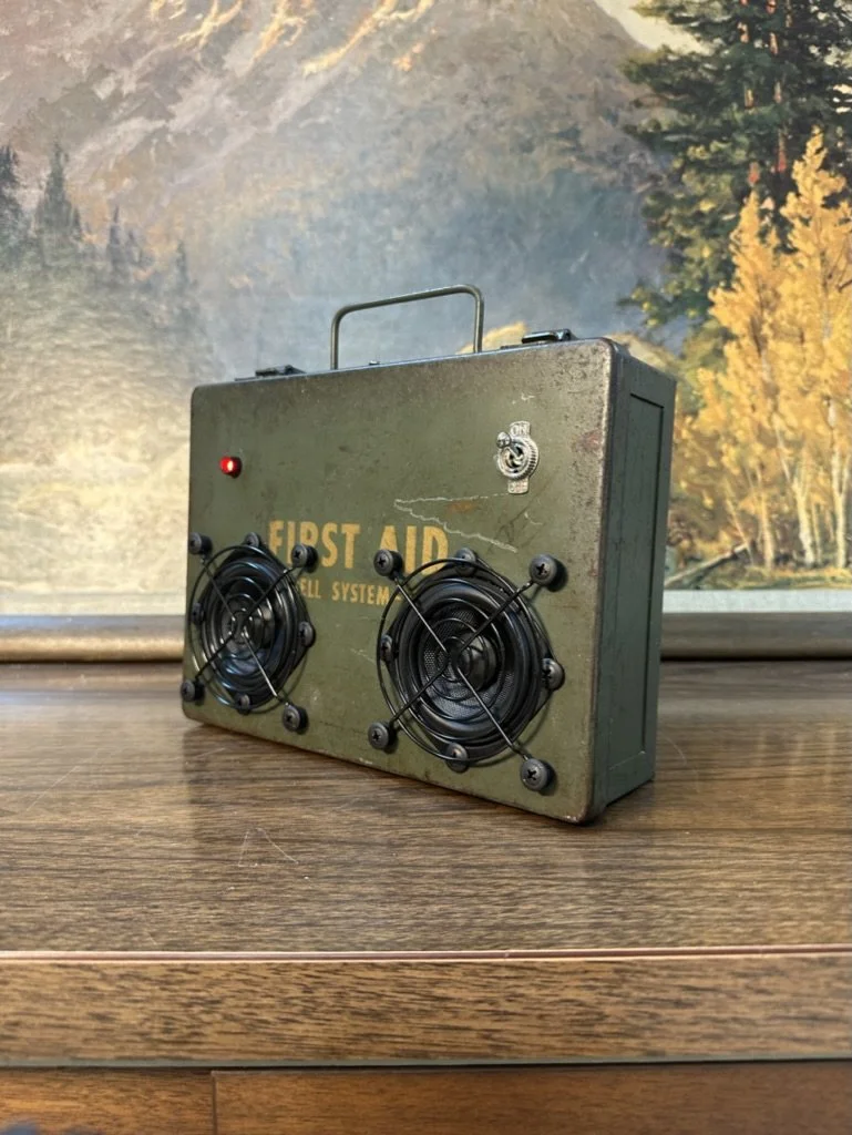 A vintage-style first aid box repurposed as a speaker, placed on a wooden surface with a nature background.