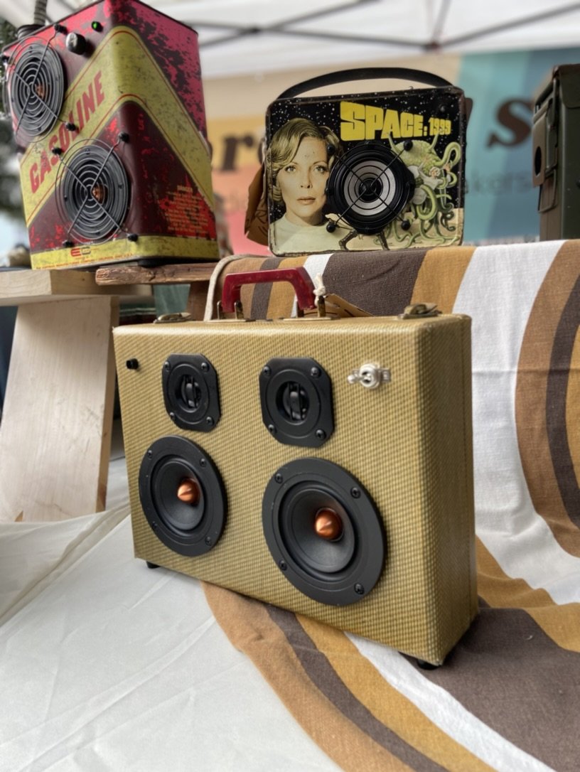 Vintage suitcase speakers displayed on a table. One has a tan fabric cover with visible speaker cones, another features a retro "Gasoline" design, and a third has a "Space: 1999" theme with a woman's face and sci-fi elements.