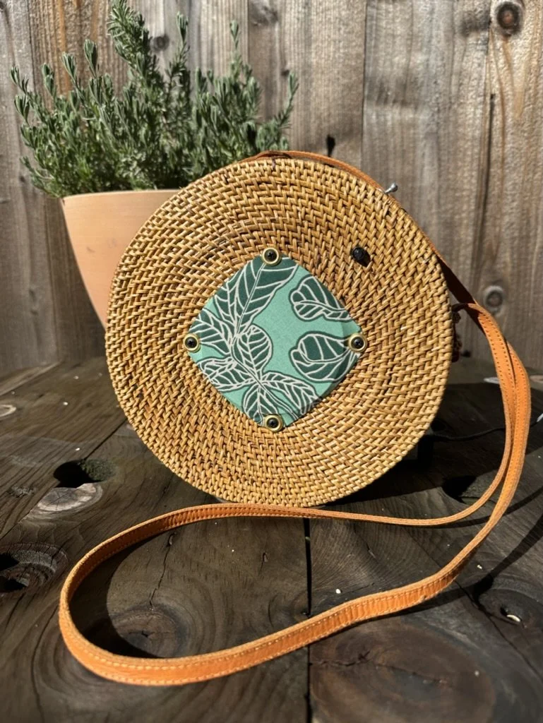 Round woven rattan bag with a leather strap and a green fabric square with a leaf pattern on a wooden surface, next to a potted plant.