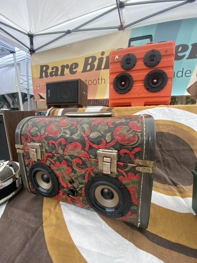 Colorful vintage-style speakers displayed on a table at a market stall. One speaker is decorated with a floral pattern, and another has an orange exterior with exposed speaker cones. A "Rare Bird" banner is visible in the background.