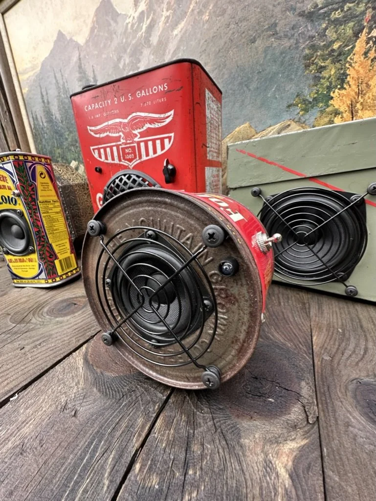 Vintage oil cans repurposed into speakers on a wooden surface, with a mountain landscape painting in the background.