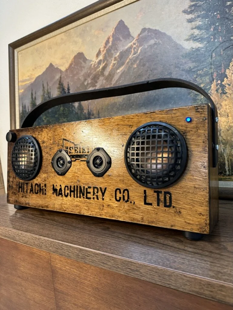 Vintage-style wooden speaker with "Hitachi Machinery Co. Ltd." written on it, placed on a wooden surface, with a mountain landscape painting in the background.