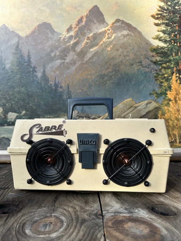 Vintage portable stereo with speakers and handle, labeled 'Sabre' and 'Umco', on a wooden table, with a mountain landscape mural in the background.