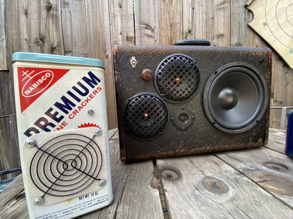 Vintage Nabisco tin repurposed as a speaker, next to an old leather-encased speaker, both on a rustic wooden table, with a wooden fence in the background.