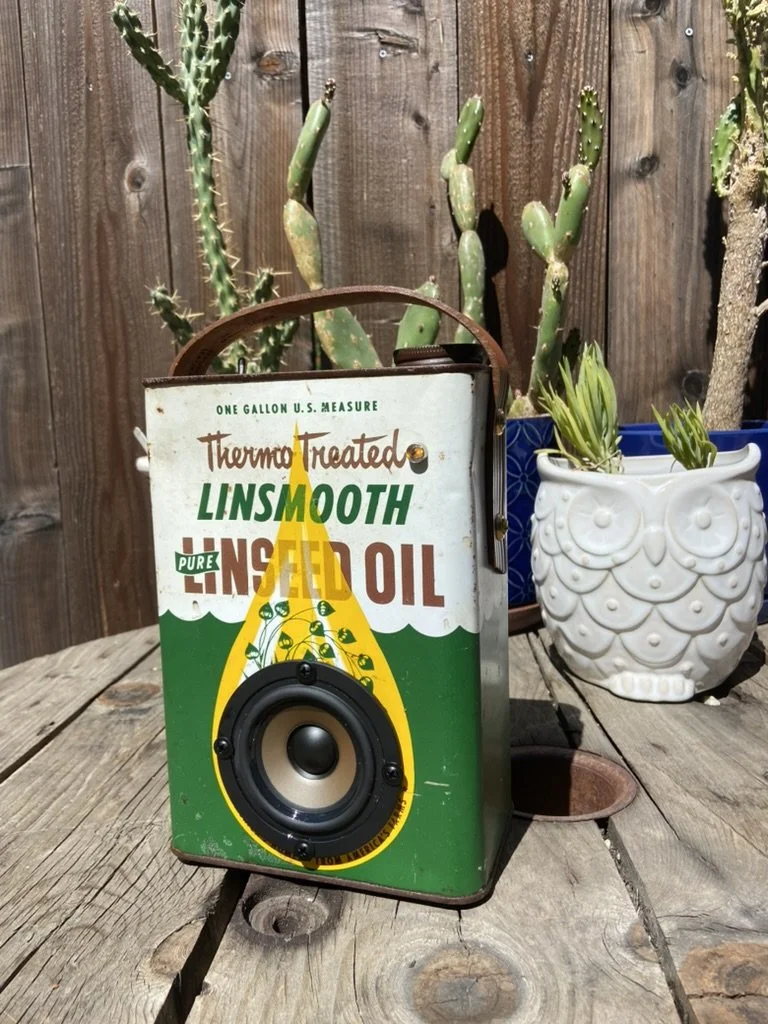 Vintage linseed oil can repurposed as a speaker on a wooden surface, surrounded by potted cacti and wooden fence in the background.