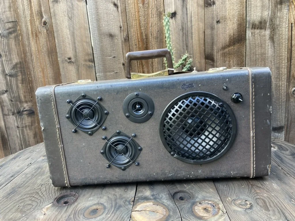 Vintage suitcase repurposed as a speaker with multiple speakers and volume knob on a wooden surface.