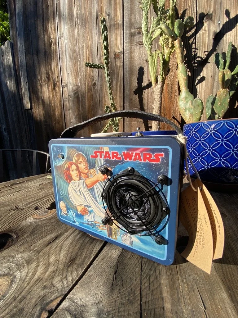 Vintage Star Wars themed tin lunchbox with retro graphic design, featuring characters from the original movie. It has a black fan attached to the front, sitting on a wooden table with cacti and a blue pot in the background.
