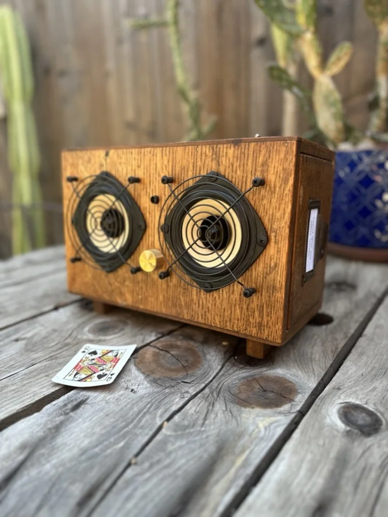 Wooden speaker with vintage style, featuring dual circular grilles, placed on a rustic wooden table with a playing card nearby. Background includes blurred outdoor plants and fence.