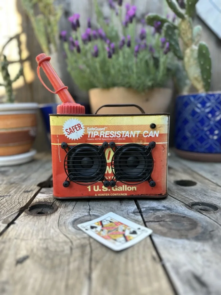 Vintage metal gas canister repurposed into a boom box with speakers, set on a wooden surface with a playing card nearby. Potted plants are in the background.