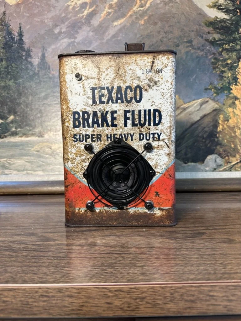 Vintage Texaco brake fluid can with a speaker on a wooden shelf, featuring a mountainous landscape painting in the background.