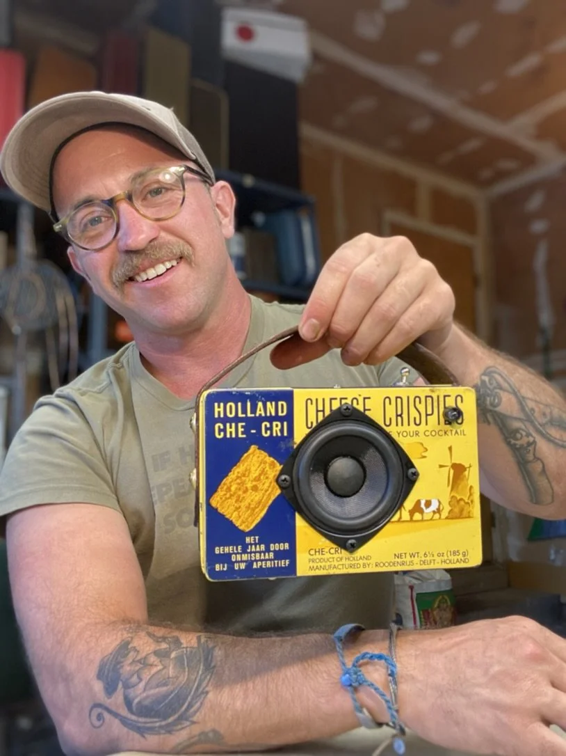 Man holding a vintage cheese crispies tin with a speaker installed, wearing glasses and a cap, tattoos on forearms, smiling in a workshop.