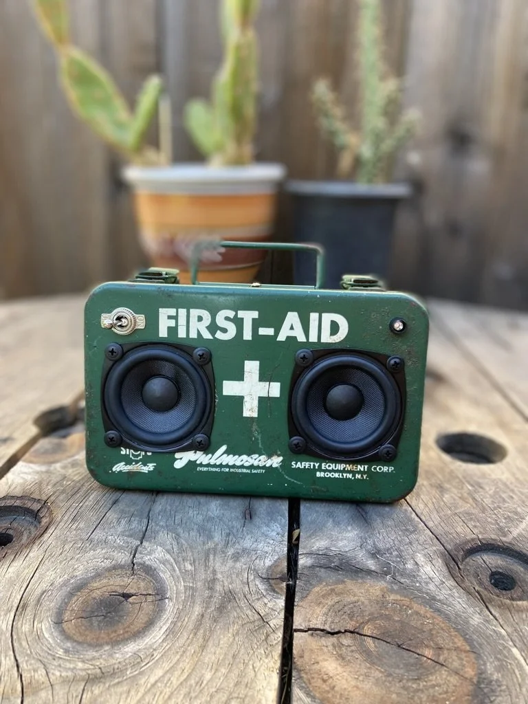 Green vintage first aid box converted into a DIY speaker with two visible speakers, placed on a wooden surface with blurred potted plants in the background.