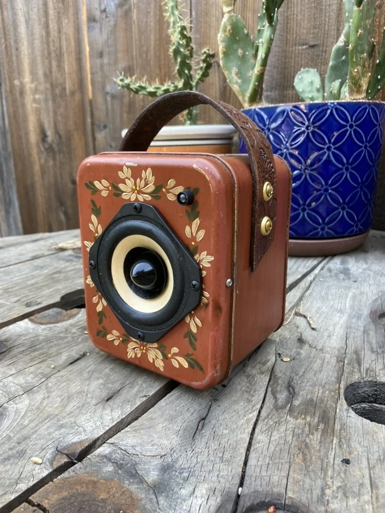 Vintage-style portable speaker with floral design and leather handle on a wooden surface, next to potted cactus plants.