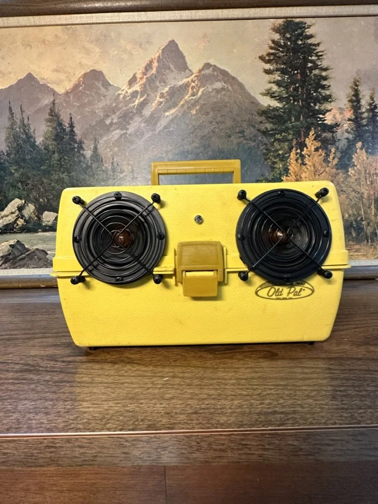 Yellow vintage Old Pal tackle box with two circular ventilation holes, set against a mountain landscape painting background. The box has a handle and a clasp on the front.