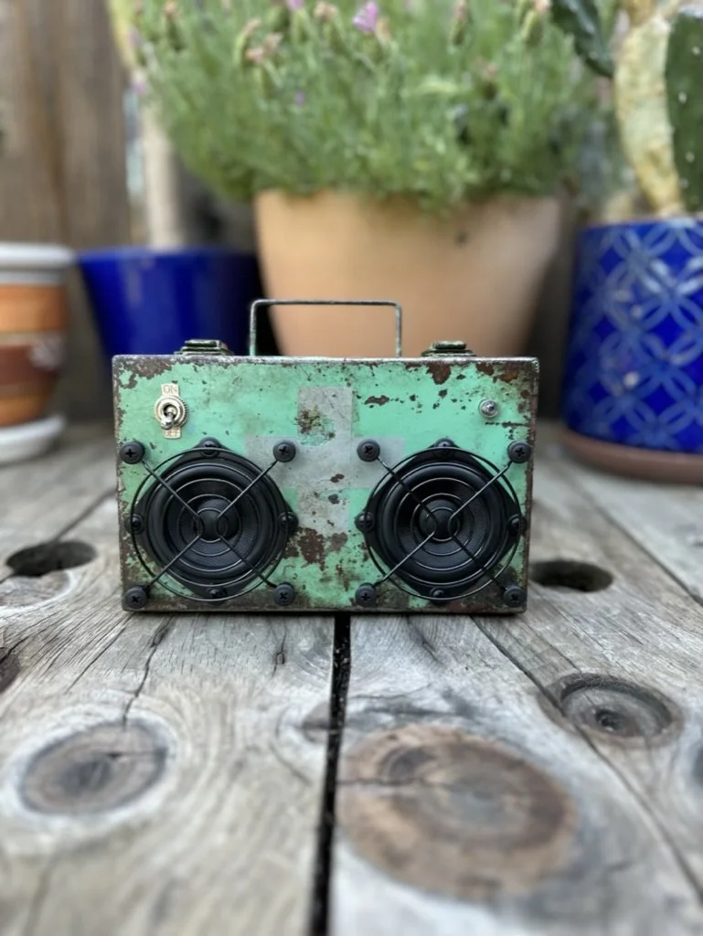 Vintage portable speaker with a rusted metal casing and dual speakers, placed on a wooden surface with potted plants in the background.