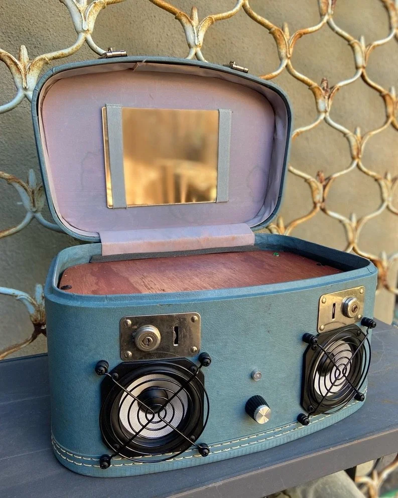 Repurposed vintage suitcase turned into a speaker system with two visible speakers, a knob, and a mirror inside the lid, placed against a decorative wall.